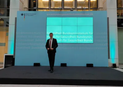 Ein Redner auf der Bühne im Bundesministerium für Gesundheit vor einer Videowand mit dem Namen des Ministeriums